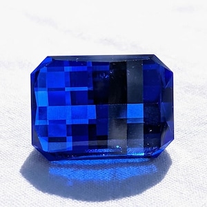 Puede incluir: Una gema rectangular azul oscuro facetada. La gema tiene un corte brillante con múltiples facetas que reflejan la luz. La piedra preciosa se encuentra sobre un fondo blanco, creando un contraste llamativo.