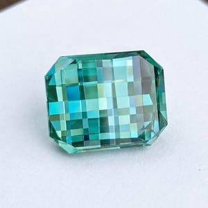 May include: A rectangular loose gemstone featuring a checkerboard cut with a vibrant teal and blue color palette. The faceted surface displays a grid-like pattern of light and dark reflections, creating a pixelated appearance across the stone.