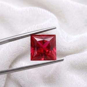May include: A square-cut, vivid red gemstone is held by silver tweezers against a white, textured background. The gemstone's facets reflect light, showcasing its clarity and colour. This image highlights the beauty and precision of the cut.