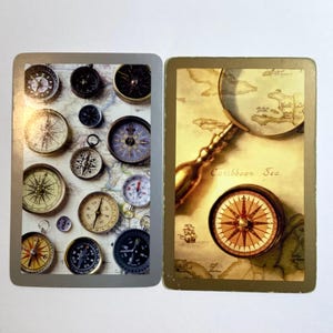 May include: Two playing cards with a nautical theme. One card features various compasses on a map background. The other card shows a compass, a magnifying glass, and a map with the text "Caribbean Sea".