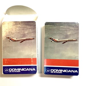 May include: Two vintage Dominicana de Aviacion playing card decks. Each deck features an image of a passenger airplane against a cloudy sky, with a red and blue stripe at the bottom. The Dominicana logo is visible.