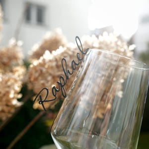 Könnte beinhalten: Ein Weinglas aus klarem Glas mit einem schwarzen Namensschild in Schreibschrift, das am Rand befestigt ist. Das Namensschild zeigt den Namen Raphael in einer dünnen, eleganten Schriftart. Das Glas ist transparent mit einer geschwungenen Form.