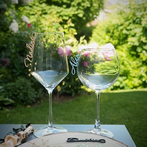 Könnte beinhalten: Zwei Weingläser aus klarem Glas mit personalisierten Namensschildern am Rand. Ein Glas hat ein hellbraunes Schild mit der Aufschrift Nadine, das andere ein hellblaues Schild mit Alex. Eine Holzscheibe zeigt weitere bunte Namensschilder in Schreibschrift.