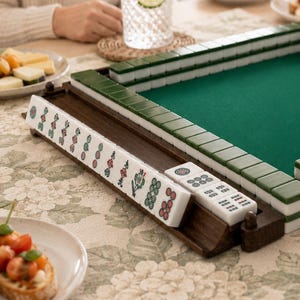 May include: A wooden Mahjong set with green and white tiles, a green felt playing surface, and a wooden tile holder. The set is on a floral patterned tablecloth, with food and drinks on the table.