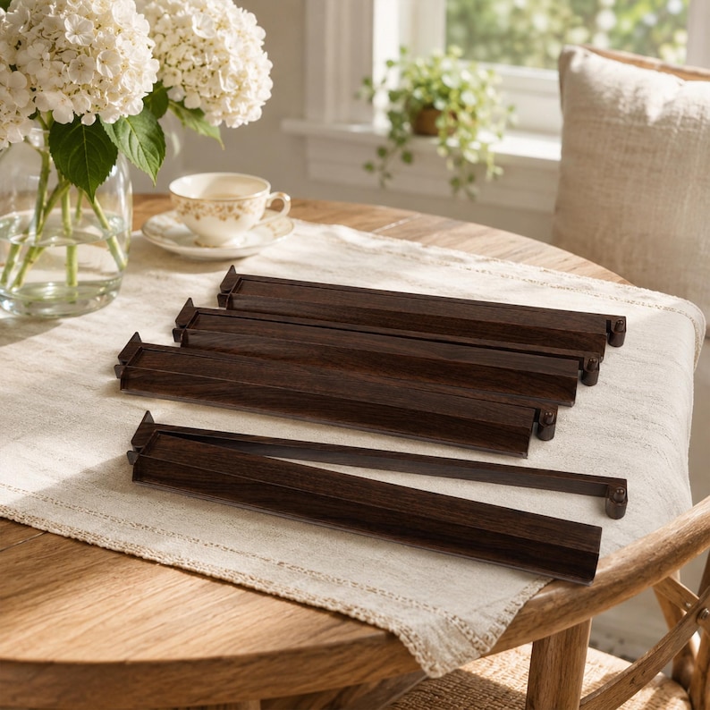 May include: Several dark brown wooden table rails are arranged on a light-colored tablecloth. The rails are rectangular with a decorative edge. A vase of white flowers and a teacup are in the background, suggesting a dining or living room setting.