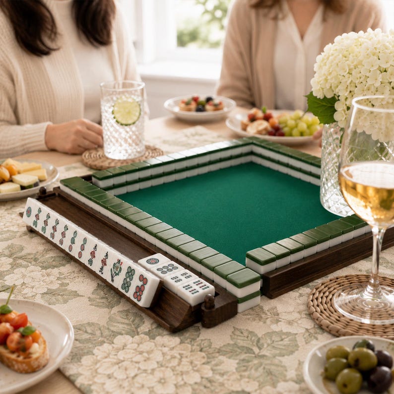May include: A wooden Mahjong set with green and white tiles, a green felt playing surface, and a wooden tile holder. The set is on a floral patterned tablecloth, with food and drinks on the table.