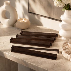 May include: A collection of dark brown wooden tile racks, likely for a board game, are arranged on a light-colored surface. The racks are rectangular with a lip on one side. Decorative vases and a lit candle are in the background.