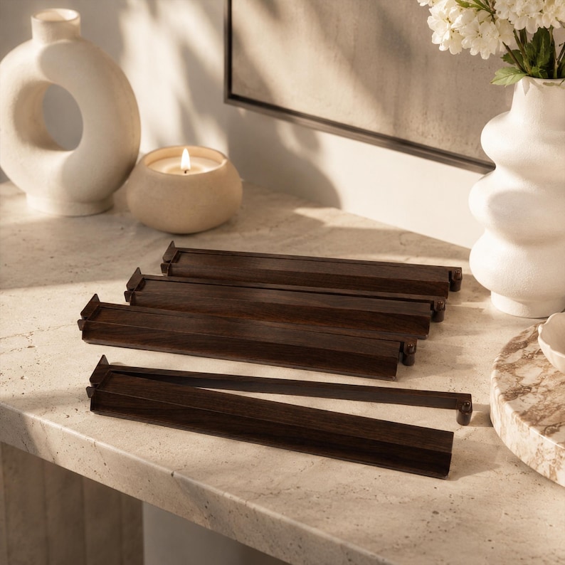 May include: A collection of dark brown wooden tile racks, likely for a board game, are arranged on a light-colored surface. The racks are rectangular with a lip on one side. Decorative vases and a lit candle are in the background.