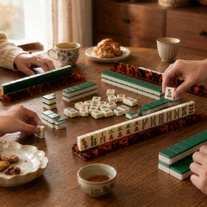 May include: A game of Mahjong in progress, with ivory and green tiles arranged on a wooden table. The tiles are set on tortoise-shell patterned racks. Teacups and snacks are also on the table, suggesting a social gathering.