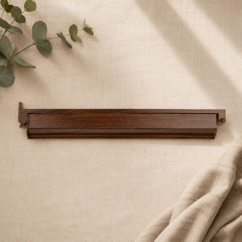 May include: A dark brown wooden shelf with a rectangular design. The shelf is mounted on a beige wall, with a sprig of eucalyptus in the upper left corner. The shelf is approximately 30 inches long.
