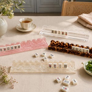 May include: A still-life image featuring several Mahjong tile racks in various colors and patterns. The racks hold white tiles with red and green floral designs. A cup of tea and a small dish of green olives are also present.