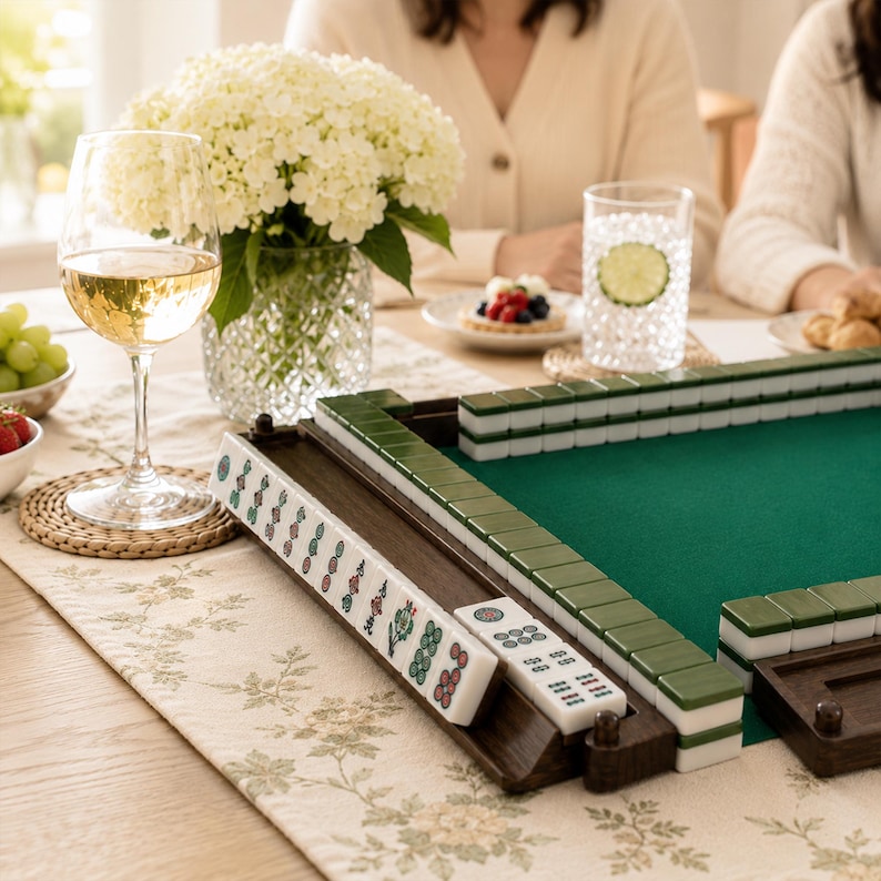May include: A wooden Mahjong set with green and white tiles is displayed on a table with a green felt playing surface. A glass of white wine, a vase of white flowers, and a glass of water with a lime slice are also on the table. The table is covered with a floral patterned runner.
