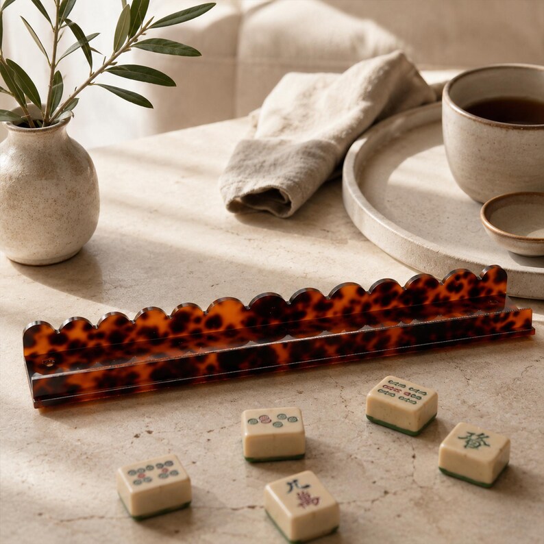May include: A tortoiseshell-patterned Mahjong tile rack with a scalloped edge, holding several ivory-colored tiles with green and red markings. The game pieces are scattered on a light-colored surface.
