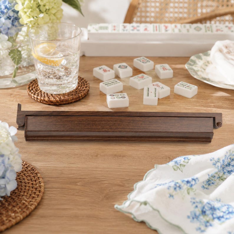 May include: A wooden mahjong tile holder with a dark brown finish, alongside white mahjong tiles with various designs. A glass of water with a lemon slice sits on a woven coaster. A floral patterned cloth and flowers are also present.