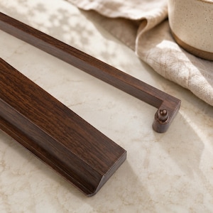May include: Close-up of two dark brown wooden picture frame pieces. The frame has a wood grain texture and a rectangular shape. One piece has a rounded knob on the end. The frame is on a light-colored surface.