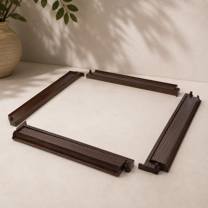 May include: Four dark brown wooden frame pieces arranged to form a square. Each piece has a grooved channel and connecting tabs. The pieces are on a light-colored surface, with a neutral background and a decorative vase.