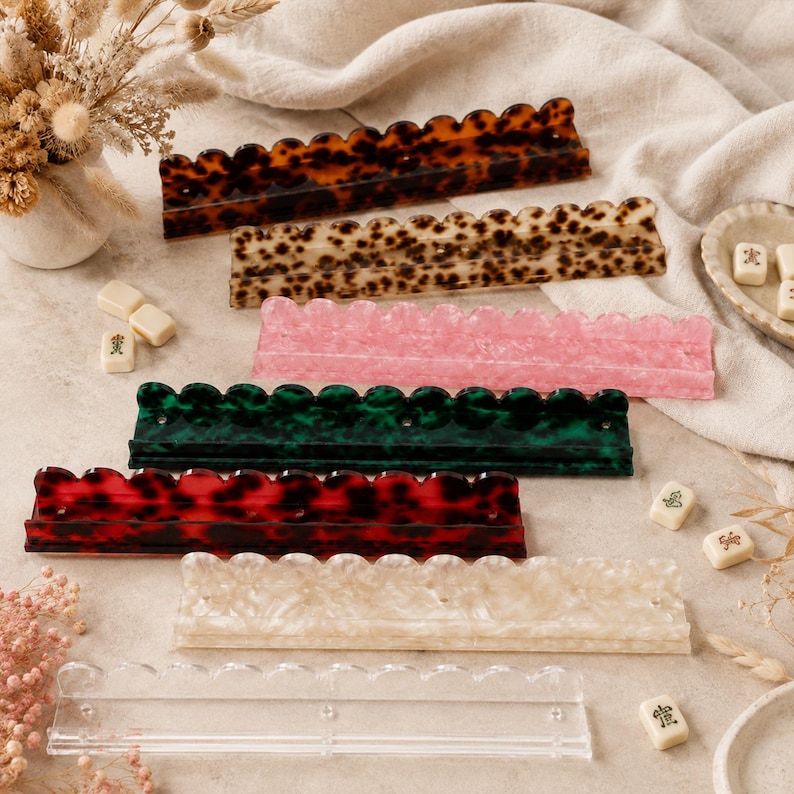May include: A collection of colorful, scalloped-edge Mahjong tile racks in various colors and patterns. The racks include tortoise shell, pink, green, red, cream, and clear acrylic. Mahjong tiles are scattered around the racks.