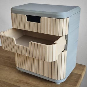 Könnte beinhalten: Ein zweischubiger Aufbewahrungsschrank mit hellblauem Äußeren und hellen, holzfarbenen Schubladenfronten. Die Schubladen haben ein einzigartiges Design mit vertikalen Holzlatten. Eine Schublade ist teilweise geöffnet und zeigt das Innere.