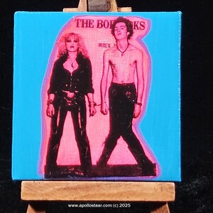 May include: A pink and blue canvas painting of two people. The people are in black clothing and are standing in front of a blue background. The text "THE BO" and "KS" are visible at the top of the painting.