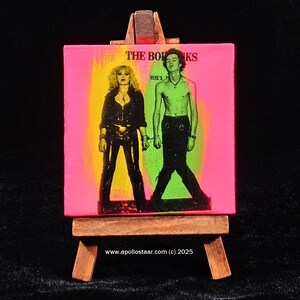 May include: A pink canvas painting on an easel with a black and white image of a band. The band is called "The Bo-R-Ks" and the text "Here's" is visible. The image is of two people, one with long black hair and the other with short black hair.