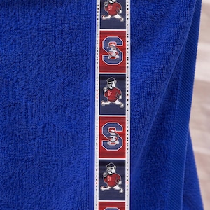 May include: A blue towel with a decorative border featuring the letter "S" and a bulldog mascot in red and white. The towel is made of a soft, absorbent material.