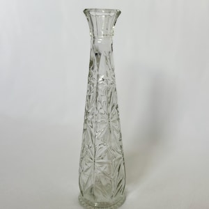 May include: A tall, clear glass bud vase with a slender, tapered silhouette. The exterior features a pressed geometric starburst pattern throughout the body, a flared rim, and a circular base with a subtle scalloped edge detail at the bottom.