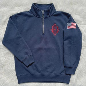 Puede incluir: Sudadera azul marino con cremallera de cuarto de cuello, monograma rojo bordado y parche de la bandera estadounidense en la manga. La sudadera tiene cuello alto y cremallera plateada.