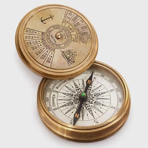 May include: A vintage brass compass with an open lid showing a calendar. The compass face displays directional markings and a needle. The lid features an anchor and ship illustration, with calendar dates. A classic navigational tool.