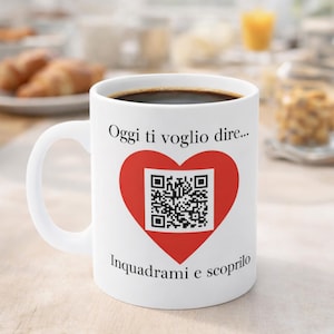 Op de afbeelding: Witte keramische koffiemok gevuld met donkere koffie. De mok heeft een rood hart met een QR-code in het midden en Italiaanse tekst. De handgreep is wit en de achtergrond toont ontbijtproducten.