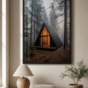 Könnte beinhalten: Ein gerahmter Wandkunstdruck, der eine schwarze A-förmige Hütte in einem dichten, nebligen Kiefernwald zeigt. Die Hütte hat große Glasfenster mit warmem Innenlicht und steht vor einer Kulisse aus hohen, dunklen Bäumen und braunem Waldboden.
