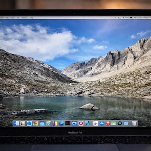 Puede incluir: Un ordenador portátil MacBook Pro muestra un paisaje de montaña con un lago bajo un cielo azul con nubes. El portátil está abierto sobre una superficie de madera. La pantalla muestra un escritorio con iconos de aplicaciones en la parte inferior.