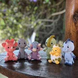 May include: A collection of six handmade crochet dinosaur figurines in various colors. The dinosaurs include a pink triceratops, a gray dinosaur, a purple dinosaur, a green dinosaur, a yellow dinosaur hatching from an egg, and a blue dinosaur.