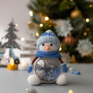 Puede incluir: Un muñeco de nieve hecho a mano con gorro, bufanda y mitones de punto azul y blanco. El muñeco de nieve tiene un cuerpo de vidrio transparente lleno de purpurina plateada y cuentas azules. Decoración navideña festiva.