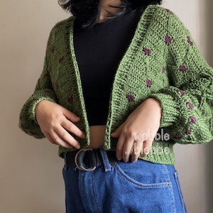 Könnte beinhalten: Eine grüne Häkeljacke mit einem floralen Muster in einem dunkleren Lilaton. Die Strickjacke hat lange Ärmel und ist vorne offen. Der Text "wobole blobbe" ist auf der rechten Seite des Bildes zu sehen.