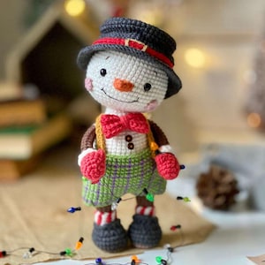 Può includere: Bambola pupazzo di neve all'uncinetto fatta a mano con cappello nero con fascia rossa, papillon rosso e pantaloncini a quadri verdi. Il pupazzo di neve ha un naso a carota, guance rosee ed è adornato da una stringa di luci natalizie colorate.