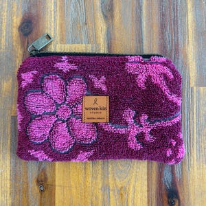 May include: A rectangular zippered pouch made of textured, deep magenta fabric featuring a repeating pattern of bright pink flowers and foliage. A small, square tan leather label is centred on the front with the text woven kin STUDIO textiles reborn.