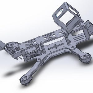 Puede incluir: Un marco de dron gris con un soporte de cámara rectangular y cuatro brazos. El marco tiene varios recortes y puntos de montaje para componentes electrónicos. El diseño parece ser una representación 3D o un modelo CAD.