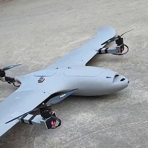 May include: A grey fixed-wing drone with a sleek aerodynamic body, featuring three visible electric motors with black propellers mounted on the wings and tail. The aircraft has a smooth, matte finish and a front-facing camera aperture.