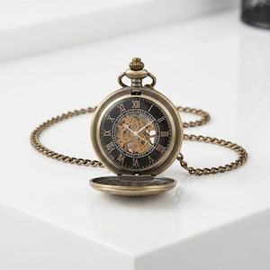 Puede incluir: Un reloj de bolsillo de estilo vintage con una caja y cadena de color bronce. La esfera del reloj es negra con números romanos y un movimiento mecánico visible. La tapa del reloj está abierta.