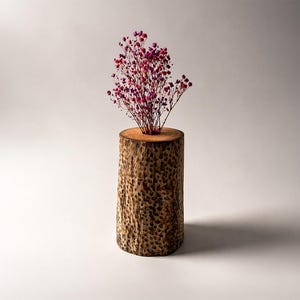 Könnte beinhalten: Ein zylindrisches Holzgefäß mit einer Höhe von etwa 20 cm, gefüllt mit kleinen rosa und lila Blüten. Das Gefäß besteht aus einem Baumstammabschnitt und zeigt eine natürliche Holzmaserung.
