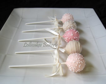 Könnte beinhalten: Eine Reihe von sechs Cake Pops auf weißen Stielen mit weißem Band, das um die Stiele gebunden ist. Die Cake Pops sind mit weißem Fondant überzogen und mit rosa und weißen Streuseln verziert. Die Cake Pops sind auf einem weißen Teller angeordnet, wobei der Text "Lollicakes Bakery" in der linken unteren Ecke sichtbar ist.