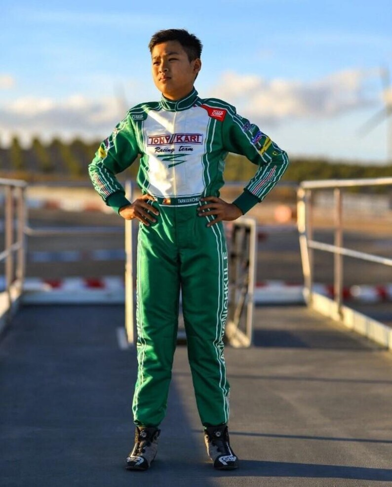 May include: A person in a green and white racing suit with the text "TONY KART Racing Team". The suit has white stripes and accents. The person is standing on a racetrack.