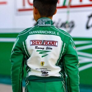 May include: Green and white racing suit with the text "Tarnvanichkul E." and "TONY KART Racing team" on the back. The suit also features the Ferrari Driver Academy logo and OMP branding. The suit has white stripes and a green collar.