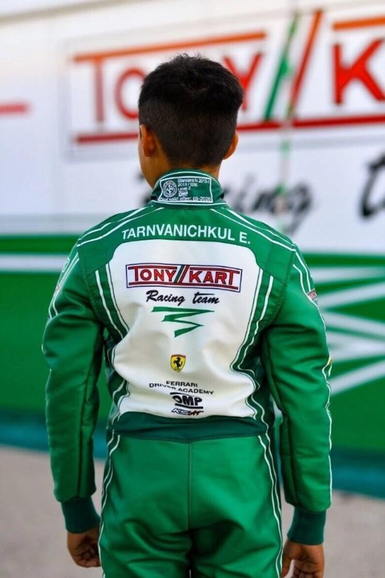 May include: Green and white racing suit with the text "Tarnvanichkul E." and "TONY KART Racing team" on the back. The suit also features the Ferrari Driver Academy logo and OMP branding. The suit has white stripes and a green collar.