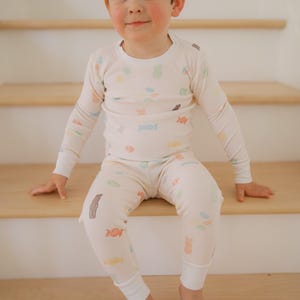 Pode incluir: Conjunto de pijama infantil creme, adornado com ilustra&ccedil;&otilde;es coloridas de doces. A parte de cima de manga comprida e as cal&ccedil;as t&ecirc;m gola redonda e punhos el&aacute;sticos. A crian&ccedil;a est&aacute; sentada em degraus de madeira.