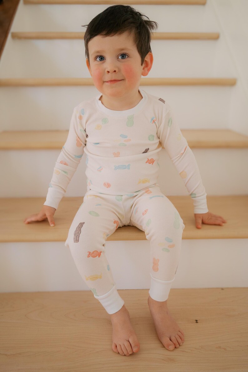 Pode incluir: Conjunto de pijama infantil creme, adornado com ilustra&ccedil;&otilde;es coloridas de doces. A parte de cima de manga comprida e as cal&ccedil;as t&ecirc;m gola redonda e punhos el&aacute;sticos. A crian&ccedil;a est&aacute; sentada em degraus de madeira.