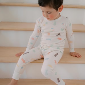 Pode incluir: Conjunto de pijama infantil de duas pe&ccedil;as em creme, adornado com um padr&atilde;o colorido de doces. A parte de cima de manga comprida e as cal&ccedil;as s&atilde;o feitas de tecido macio. A crian&ccedil;a est&aacute; sentada em degraus de madeira.