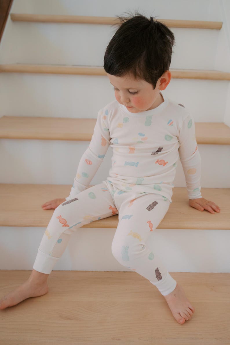 Pode incluir: Conjunto de pijama infantil de duas pe&ccedil;as em creme, adornado com um padr&atilde;o colorido de doces. A parte de cima de manga comprida e as cal&ccedil;as s&atilde;o feitas de tecido macio. A crian&ccedil;a est&aacute; sentada em degraus de madeira.