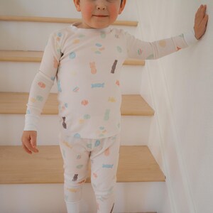 Pode incluir: Uma crian&ccedil;a vestindo um conjunto de pijama creme com um padr&atilde;o repetido de doces e coelhos da P&aacute;scoa em cores pastel. A parte superior de manga comprida e as cal&ccedil;as t&ecirc;m punhos brancos. A crian&ccedil;a est&aacute; em p&eacute; em uma escada de madeira.