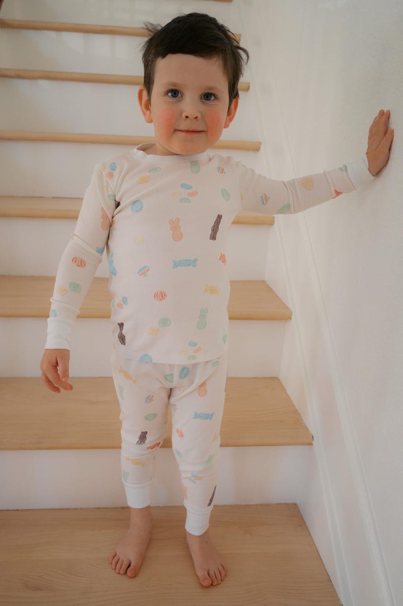 Pode incluir: Uma crian&ccedil;a vestindo um conjunto de pijama creme com um padr&atilde;o repetido de doces e coelhos da P&aacute;scoa em cores pastel. A parte superior de manga comprida e as cal&ccedil;as t&ecirc;m punhos brancos. A crian&ccedil;a est&aacute; em p&eacute; em uma escada de madeira.